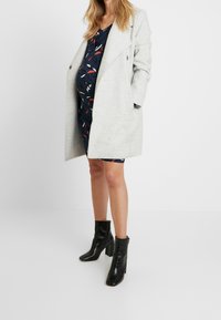 Grå ullkappa med en mjuk textur, sidofickor, lagd över en marinblå klänning med ett färgglatt fjädertryck. Svarta högklackade ankelstövlar.