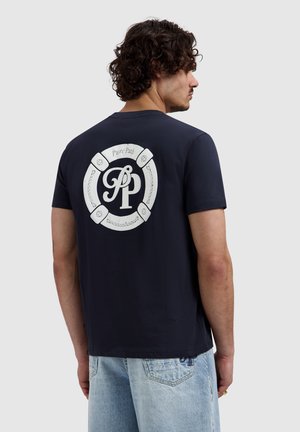 Man met krullend haar, gekleed in een navy T-shirt met een wit rond "PP Pure Path" logo op de rug en lichtblauwe spijkerbroek, kijkend naar rechts.