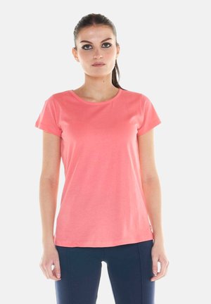 Frau mit dunklem Haar im Pferdeschwanz, trägt ein korallenrosa kurzärmeliges T-Shirt und dunkle Hosen, steht vor einem schlichten hellen Hintergrund.