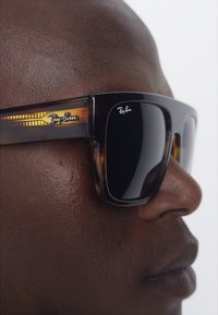 Ochelari de soare Ray-Ban cu ramă din țestoasă, lentile închise la culoare și un logo distinct pe bratul ochelarilor. Au o formă rectangulară și un finisaj lucios.