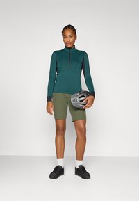 Moteris stovi su pasitikėjimu, vilkinti mėtų spalvos zip-up ilgomis rankovėmis marškinėlius ir alyvuogių spalvos šortus, laikydama pilką šalmą, su neutraliu fonu.