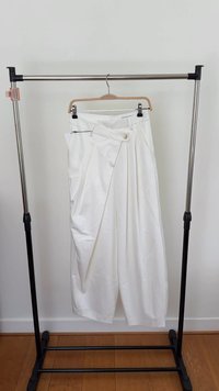 Białe, szerokie spodnie na wieszaku, z przednim zapięciem na guzik, detalami w formie zakładek oraz gładką teksturą materiału. Odpowiednie na różne okazje.