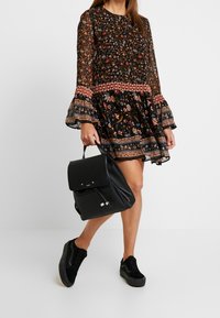 Zaino in pelle nera con chiusura a pattina e tasca frontale, abbinato a un vestito a maniche lunghe con motivo floreale e scarpe da ginnastica nere.