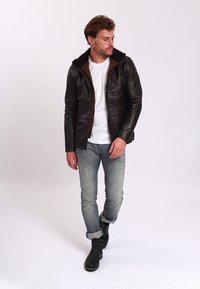 Brun läderjacka med huva och kontrasterande brun kant, bärs över en vit T-shirt. Jeans är ljusblå med slitna detaljer. Svarta kängor.