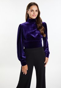 Top din catifea violet cu mâneci bufante lungi și guler înalt, combinat cu pantaloni largi negri. Textură netedă, talie ajustată și design elegant.