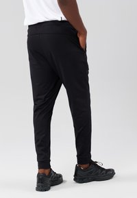 Zwarte sportbroek met een taps toelopende pasvorm, elastische tailleband en zijzakken, gecombineerd met zwarte sportschoenen op een lichtgrijze achtergrond.