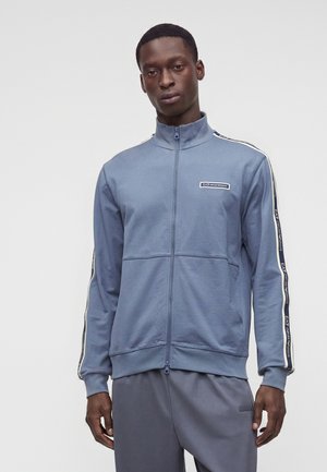 Homme portant une veste de sport bleue zippée avec un logo sur la poitrine et des bandes de marque sur les manches, associée à un pantalon bleu assorti.