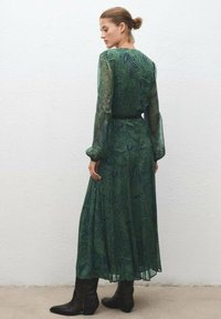 Largo vestido verde con tela transparente y fluida que presenta intrincados patrones de paisley. Mangas largas, cinturón en la cintura y botas negras hasta la rodilla.