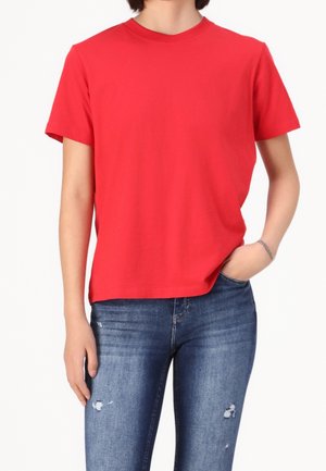 Persona vistiendo camiseta lisa roja de manga corta y jeans azules desgastados, de pie con una mano en el bolsillo contra un fondo blanco.