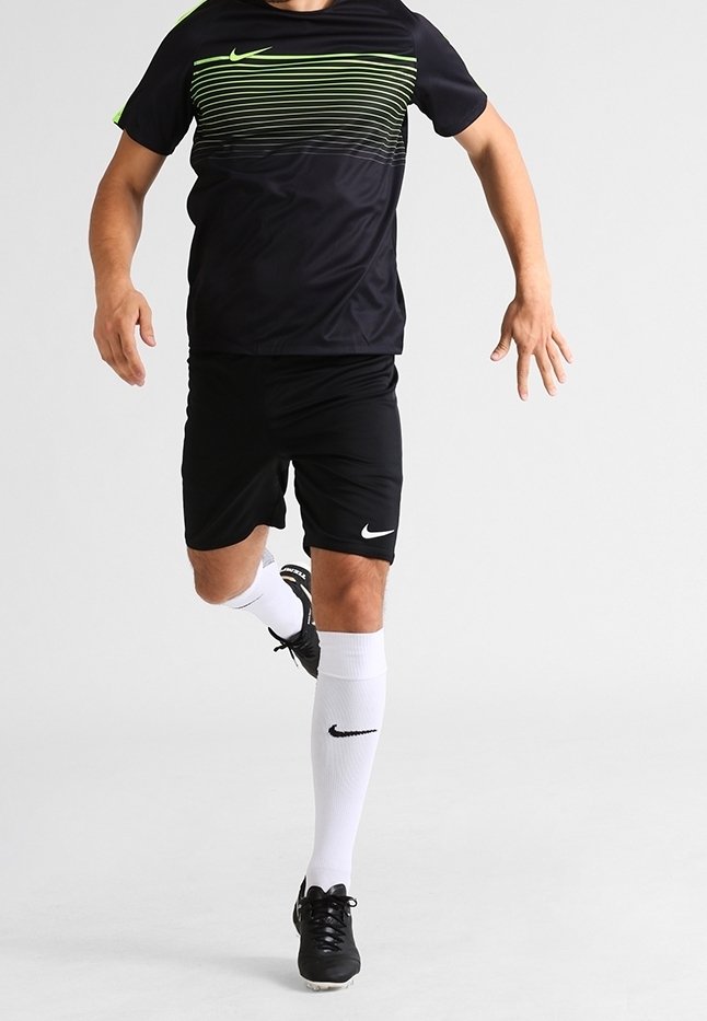 Athlète courant portant des vêtements de sport Nike noirs et verts, un short noir, des chaussettes blanches montantes et des crampons noirs sur fond gris.