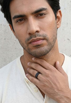 Junger Mann mit leichtem Stoppelbart, der ein weißes Hemd trägt und mit einer Hand seinen Hals berührt, an der ein schwarzer Ring am Mittelfinger sitzt.