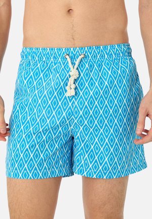 Short de bain bleu avec motif de losanges blancs et cordon blanc porté par une personne, montrant la taille et le haut des jambes.