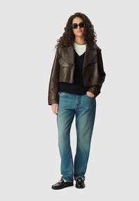 Bruine leren korte jas met zilveren details, zwarte trui, lichtblauwe spijkerbroek en zwarte schoenen; model draagt een zonnebril.