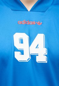 Μπλε αθλητική φανέλα με κόκκινο λογότυπο adidas πάνω από μεγάλο λευκό αριθμό 94 στο στήθος, με λαιμόκοψη σε σχήμα V.