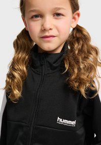 Czarna kurtka z zamkiem błyskawicznym, z teksturowaną górną częścią. Posiada białe logo marki "hummel PERFORMANCE" na klatce piersiowej.