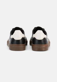 Sneakers in pelle bianca e nera con superficie strutturata, suola in gomma marrone scuro e dettagli bianchi smerlati sul tallone.