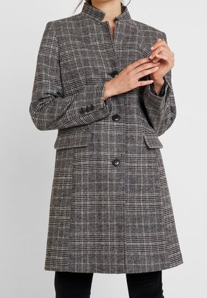 Woman wearing a knee-length gray plaid coat with black buttons and flap pockets, hands delicately clasped near chest.