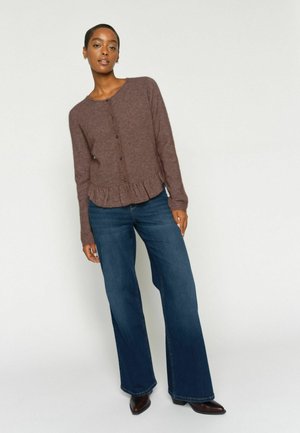 Femme portant un cardigan marron boutonné avec un ourlet à volants, un jean bleu à jambes larges, et des bottines pointues marron foncé, debout devant un fond blanc.