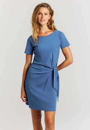 Jonge vrouw met blond haar, gekleed in een blauwe jurk met korte mouwen en een strik aan de zijkant, staand voor een effen lichte achtergrond.