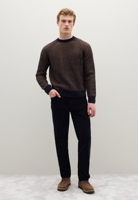 Braun-navy gestreifter Pullover mit rundem Ausschnitt, gerippten Bündchen und Saum, kombiniert mit dunkler Hose und braunen Wildlederschuhen vor hellem Hintergrund.