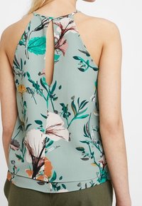 Top de halter con estampado floral en tejido verde claro, con un escote en forma de ojal, que presenta acentos florales multicolores y diseños de hojas.