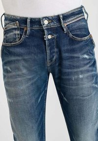 Jeans de denim en azul oscuro con un acabado desgastado, que presentan una cremallera, dos bolsillos frontales y un pequeño bolsillo para monedas en el lado derecho.