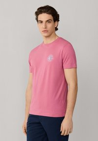 Giovane uomo con capelli scuri e ondulati che indossa una t-shirt rosa a maniche corte con un piccolo logo bianco sul petto e pantaloni blu scuro.