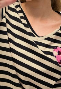 Černá a béžová pruhovaná látka s výstřihem do V. Obsahuje růžovou texturovanou nášivku ve tvaru srdce, která zdůrazňuje kontrast barev a textur.