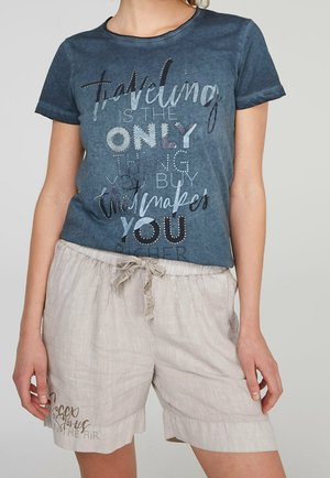 Femme portant un t-shirt bleu avec une citation sur le voyage et un short beige clair avec cordon de serrage et détail de texte sur la jambe gauche.