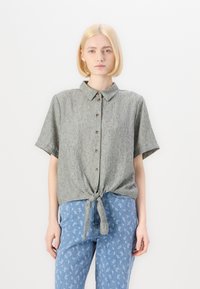 Camicia a maniche corte con colletto in tessuto grigio testurizzato, dotata di chiusura con bottoni sul davanti e dettaglio con cravatta annodata in vita, abbinata a jeans blu con motivo.