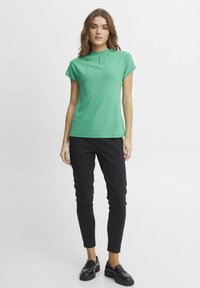 Top verde cu maneci scurte, cu guler înalt și detalii de pliu, asociat cu pantaloni negri slim-fit și loafers negri. Textură netedă pe tot parcursul.