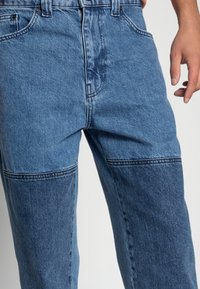 Světle modré denimové džíny s dvoubarevným designem a tmavšími denimovými panely. Standardní styl s pěti kapsami a viditelné poutka na opasek.