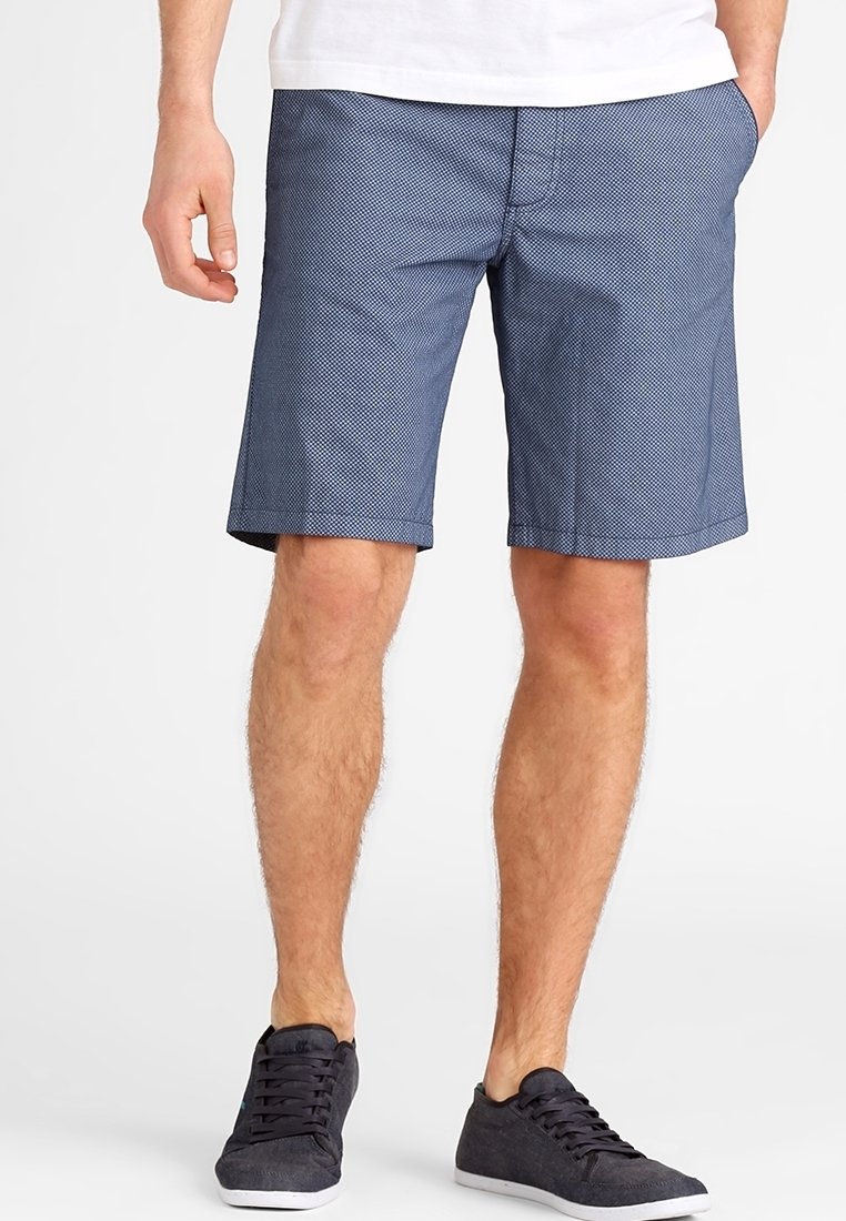 Homme portant un short bleu à motifs longueur genou, chemise blanche et baskets noires, les mains dans les poches, debout sur fond blanc.