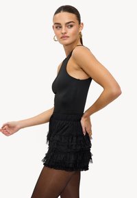 Vrouw in een zwarte mouwloze top en een gelaagde minirok met ruffles, met zwarte panty, gouden oorringen en ringen, poseert zijwaarts op een witte achtergrond.