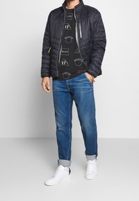 Chaqueta acolchada negra con un alto cuello, llevada sobre una camiseta gráfica negra con patrones geométricos blancos, combinada con jeans azules y zapatillas blancas.