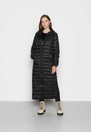 Black puffer coat with a long, quilted design, two front pockets, and a zipper closure; paired with cream-colored lace-up boots.