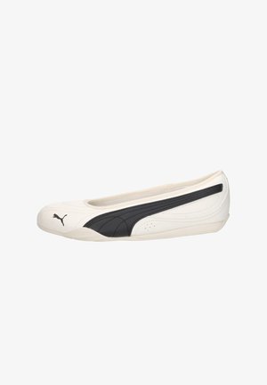 Zapato plano blanco Puma con perfil bajo, superficie lisa y una franja negra con el logo de Puma a lo largo del lateral y la puntera.