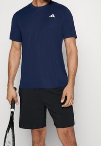 Marinblå träningströja i slätt tyg, med en vit logotyp på övre vänstra delen. Buren med svarta shorts och håller en tennisracket.