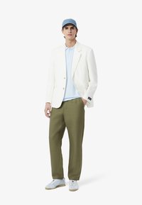 Witte blazer over een lichtblauw polo, olijfgroene broek en witte sneakers. Blauwe pet, ontspannen pasvorm, soepele stof en casual stijl.