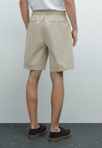 Beige katoenen shorts met elastische tailleband, zij- en achterzakken, en een knielengte, gecombineerd met zwarte suède schoenen.