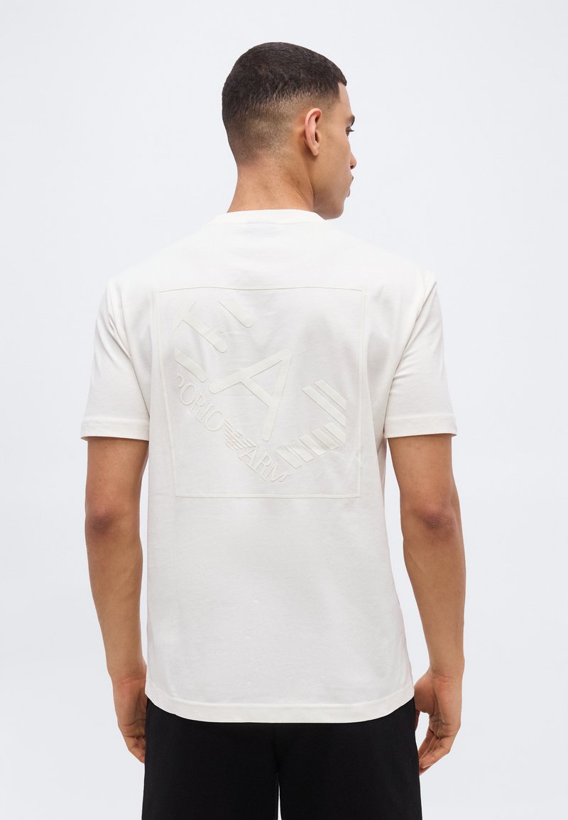 Άνδρας που φοράει λευκό t-shirt με κοντό μανίκι και ανάγλυφο λογότυπο "EA7 Emporio Armani" στην πλάτη, με την πλάτη γυρισμένη προς την κάμερα.