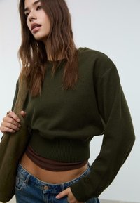 Giovane donna con lunghi capelli castani, che indossa un maglione corto verde oliva sopra una maglietta marrone, jeans blu e porta una borsa a tracolla verde oliva.