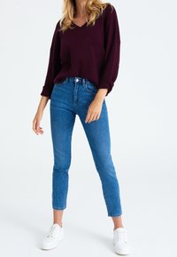 Nainen, jolla on päällä tumman violetti neule, siniset tiukat farkut ja valkoiset lenkkarit, seisoo valkoista taustaa vasten.