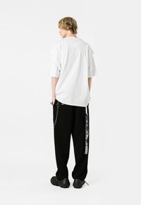 Camiseta blanca de gran tamaño con mangas cortas y un bolsillo en la parte posterior, combinada con pantalones negros holgados que presentan una cadena y una marca lateral.