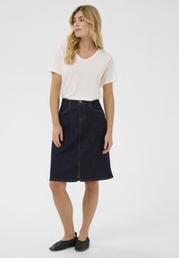 Tamsiai mėlynas džinso sijonas iki kelių su priekyje užsegamais mygtukais, dviem priekinėmis kišenėmis ir kontrastingomis siūlėmis. Derinamas su šviesia v formos iškirptės palaidine.