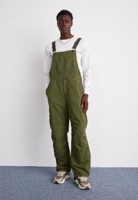 Olivgrüne Baumwoll-Latzhose mit verstellbaren Trägern, Fronttasche, weiter Passform und Seitentaschen, kombiniert mit einem weißen Langarmshirt und Sneakers.