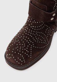 Stivale alla caviglia in suede marrone con borchie argentate a forma di spirale, dotato di una cintura regolabile e suola liscia.