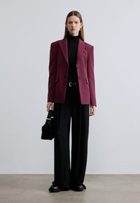 Bordeauxrode blazer met scherpe revers, zwarte coltrui, wijde zwarte broek en stevige zwarte schoenen. Houdt een kleine zwarte tas vast. Minimalistische setting.