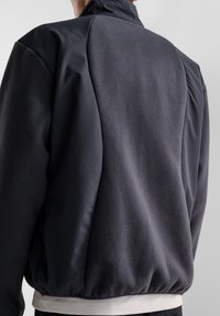 Mörkgrå jacka med en blandning av texturerad fleece och slät tyg, med hög krage och elastisk nederkant, synliga sömdetaljer.