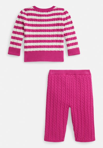 Ružový a biely pruhovaný pletený sveter s rebrovanými manžetami v kombinácii s ružovými pletenými nohavicami s copánkovým vzorom na bielom pozadí.